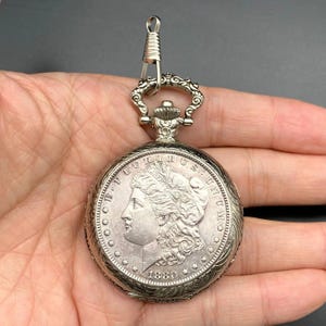 May include: Silver pocket watch with an ornate design. The watch face features a detailed portrait and the year 1880. The watch has a decorative top with a clasp for attaching to a chain. The watch is round and has a detailed edge.