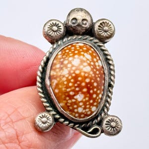 May include: A silver-toned ring featuring an oval, speckled, orange-brown stone. The ring has a decorative rope-like border and is adorned with small, circular embellishments. The design includes a skull-like element at the top.