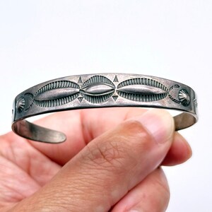 May include: A silver cuff bracelet with an engraved geometric design. The bracelet features oval and triangular patterns, with a curved shape. The bracelet is held in a hand against a white background.