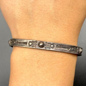 以下が含まれることがあります： 彫刻が施されたシルバーカラーのカフブレスレット。ブレスレットには、矢、花のモチーフ、幾何学模様の繰り返し模様があります。ブレスレットの幅は約1.3cmです。