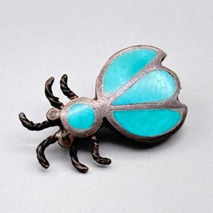 May include: A silver-toned insect brooch with turquoise-colored accents. The brooch features a detailed design with six legs and two wings. The wings and body have turquoise inlays, creating a striking contrast against the silver metal.