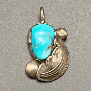 May include: A silver pendant featuring a vibrant turquoise stone. The pendant has a teardrop shape with a detailed feather and a small ball on the side. The turquoise stone is a bright blue color with brown veining.