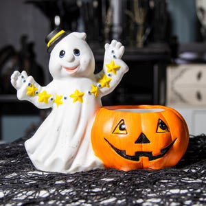 May include: A white ceramic ghost figure with a black hat and yellow star accents, next to an orange jack-o'-lantern. The ghost has a smiling face and outstretched arms. The jack-o'-lantern has a carved face. Halloween decor.