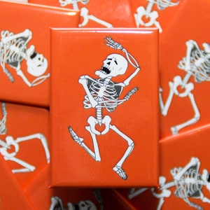 May include: A set of orange square magnets with a white skeleton illustration on each. The skeleton is in a dancing pose with its arms raised and its legs spread apart. The skeleton is smiling with its mouth open.