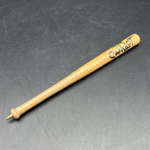 May include: A wooden pen shaped like a baseball bat, with the word "CAMOT" printed in black on the barrel. The pen has a gold-colored tip and a light brown finish.