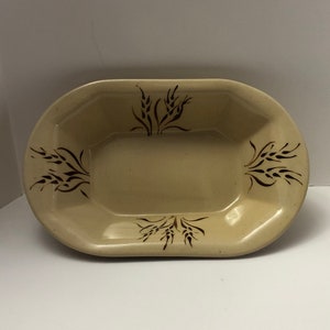 May include: A tan-colored, oval-shaped ceramic serving dish. The dish features a rectangular center and is decorated with dark brown wheat stalk designs on the rim. The dish is empty and sits on a white surface.