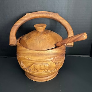 May include: A wooden sugar bowl with a lid and handle, featuring carved animal and landscape designs. The bowl includes a small wooden spoon. The bowl is brown and sits on a black surface.