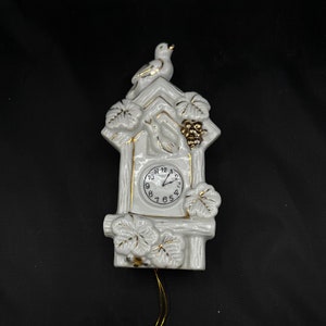 May include: White ceramic cuckoo clock with gold accents. The clock features a bird on top, grape leaf and cluster details, and a pendulum. The clock face has black numbers and hands. A decorative piece for a home.