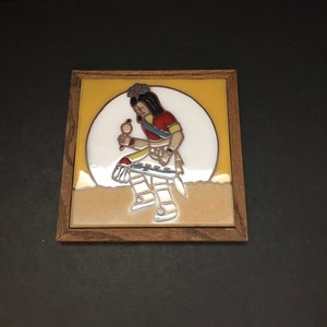 May include: A framed tile featuring a Native American figure in traditional attire. The figure is depicted in a stylized pose, holding an object, with a yellow and white background. The tile is set within a wooden frame.