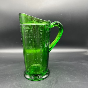 May include: A vintage green glass measuring pitcher with a handle and a spout. The pitcher has measurement markings for flour, sugar, and liquid. The base is round, and the handle is curved.