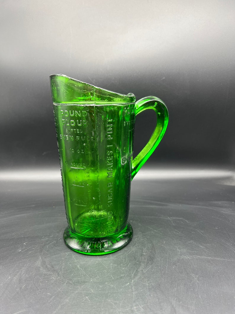 May include: A vintage green glass measuring pitcher with a handle and a spout. The pitcher has measurement markings for flour, sugar, and liquid. The base is round, and the handle is curved.