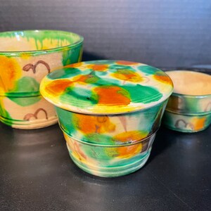 May include: Three ceramic pots with a hand-painted design. The pots are in shades of green, orange, and tan. The largest pot is a bowl shape, the middle pot is a lidded jar, and the smallest is a small bowl.