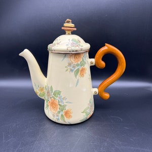 May include: Cream-colored enamelware coffee pot with a floral design featuring orange and blue flowers. The pot has a wooden handle and a matching lid with a decorative finial. The spout is curved, and the pot has a classic, vintage aesthetic.