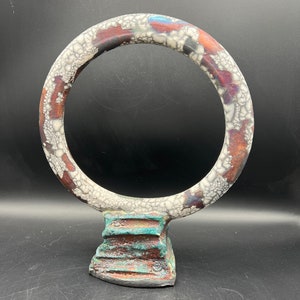 May include: A ceramic sculpture featuring a circular form atop a stepped base. The ring has a textured surface with white, brown, and blue accents. The base has a turquoise and brown color scheme, creating a unique artistic piece.