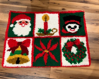 May include: A rectangular Christmas rug with six squares featuring festive designs. Santa Claus, a candle, and a snowman are in the top row. The bottom row includes a bell, a poinsettia, and a wreath. The rug has a red border.