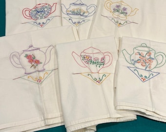 May include: White tea towels with embroidered teapots in various colors. Each towel features a different teapot design with floral accents and decorative stitching. The towels are folded and arranged on a teal surface.