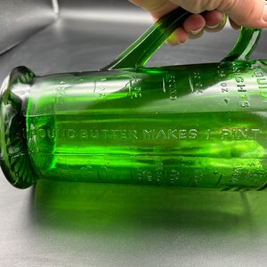 May include: A green glass measuring pitcher with a handle. The pitcher has measurement markings in ounces and cups. The text "1 POUND BUTTER MAKES 1 PINT" is visible. The pitcher is made of clear green glass.