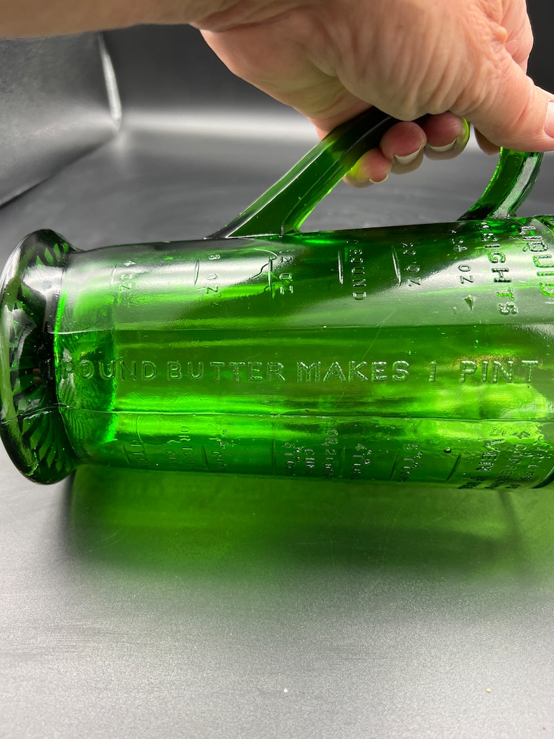 May include: A green glass measuring pitcher with a handle. The pitcher has measurement markings in ounces and cups. The text "1 POUND BUTTER MAKES 1 PINT" is visible. The pitcher is made of clear green glass.