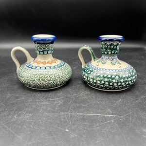 May include: Two ceramic candle holders with handles. The candle holders are round with a bulbous body and a narrow neck. The candle holders are decorated with blue, green, and orange floral patterns. The rims are blue.