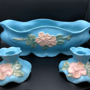 Puede incluir: Un centro de mesa de cerámica azul claro y dos candelabros a juego. El conjunto presenta diseños florales en relieve en rosa y verde. El cuenco tiene un borde festoneado y los candelabros tienen una base en forma de flor.