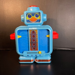 May include: A vintage blue toy robot named "ALPHIE" with a red top and arms. The robot has a smiling face, a series of red lights, and a number 7. The robot's body has a circuit board design.