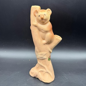 May include: A ceramic koala bear figurine clinging to a tree branch. The figurine is primarily beige with brown accents, and features a small opening at the top. A small floral design is visible on the tree trunk.