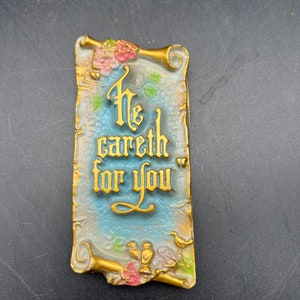 May include: Rectangular plaque with the words "He careth for you" in gold lettering. The plaque has a light blue background with gold scrollwork and floral accents in pink and green. The plaque has a vintage aesthetic.
