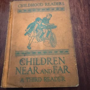 May include: A vintage book titled "CHILDREN NEAR AND FAR A THIRD READER." The cover is tan with green text and a teal illustration of two children on a bicycle. The book is part of the "CHILDHOOD READERS" series.