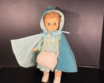May include: A vintage doll wearing a light blue dress, matching cape, and hood. The doll has blonde hair and blue eyes, with a white fluffy purse. The cape has a patterned design. The doll is wearing white shoes.