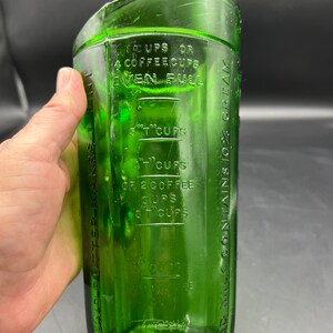May include: A vintage green glass measuring cup with embossed measurement markings. The cup has a rectangular shape with a curved top and a wide base. The markings include measurements in cups and coffee cups.