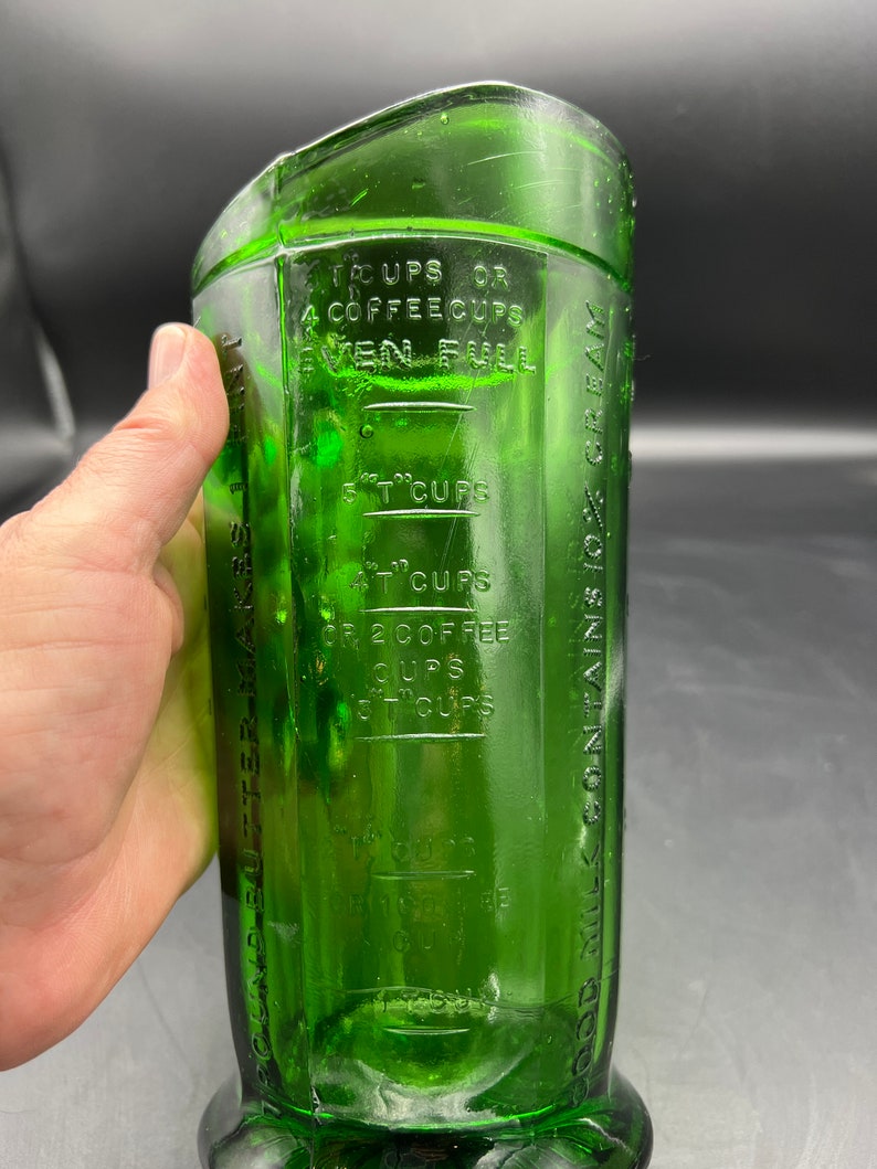 May include: A vintage green glass measuring cup with embossed measurement markings. The cup has a rectangular shape with a curved top and a wide base. The markings include measurements in cups and coffee cups.