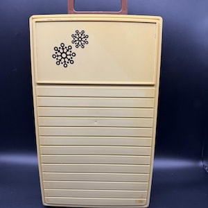 May include: A vintage, rectangular, beige plastic carrying case with a brown handle. The top section features two black snowflake designs. The lower section has horizontal, ridged detailing. A retro-style storage container.