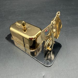 May include: A gold-colored, vintage-style desk accessory, possibly a letter opener or paper cutter, with a rectangular base and a hinged mechanism. The accessory has a shiny, reflective surface.