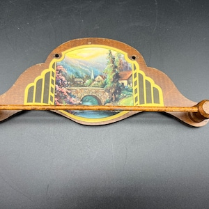 May include: Wooden towel rack with a scenic landscape print. The print features a stone bridge, a church, and a river. The rack has a brown finish with decorative accents and a wooden dowel for hanging towels.