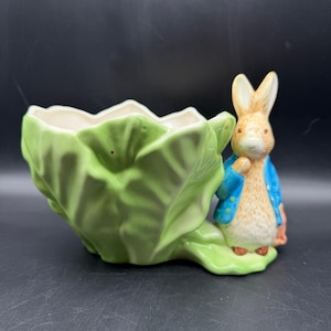 May include: A ceramic planter shaped like a green cabbage with a beige interior. A small figurine of a rabbit wearing a blue jacket and holding a small orange bag is next to the cabbage. The planter is on a dark surface.