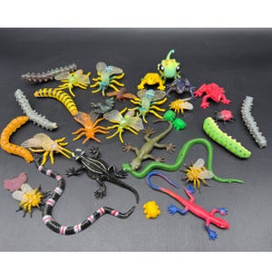 May include: A collection of colorful plastic toy insects and reptiles, including bees, lizards, caterpillars, and a snake. The toys are in various colors such as green, yellow, red, and black, and are arranged on a dark surface.