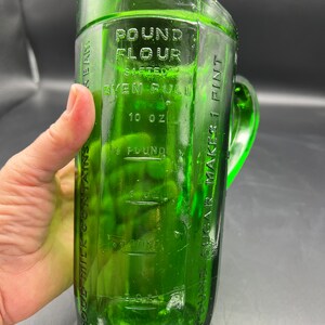 May include: A vintage green glass measuring pitcher with a handle. The pitcher has embossed markings for flour, sugar, and milk, with measurements in ounces and pounds. The text on the pitcher reads "Pound Flour Sifted Even Full" and "10 oz".