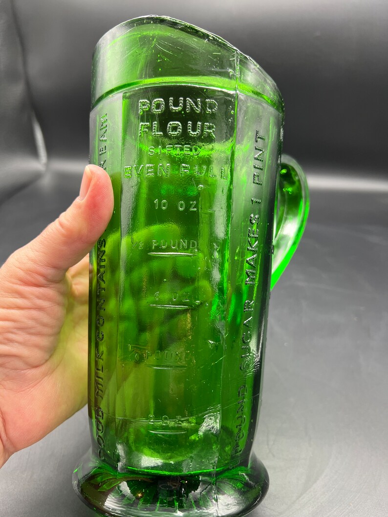 May include: A vintage green glass measuring pitcher with a handle. The pitcher has embossed markings for flour, sugar, and milk, with measurements in ounces and pounds. The text on the pitcher reads "Pound Flour Sifted Even Full" and "10 oz".