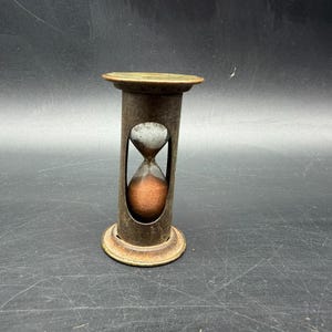 May include: A vintage hourglass with a bronze-colored metal frame. The hourglass contains brown sand and has a classic, antique design. The base and top are circular, and the glass bulb is visible through the frame.
