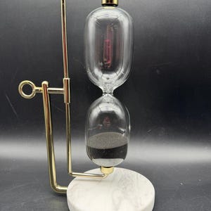May include: A rotating hourglass featuring two clear glass bulbs filled with fine black sand. The hourglass is supported by a polished gold-tone metal frame and mounted on a circular white marble base with subtle grey veining throughout the stone.