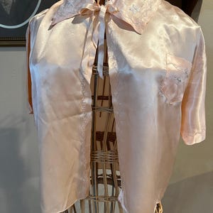 May include: A light pink satin top with short sleeves and a tie closure. The top has a collared neckline and a pocket on the left side. Delicate white embroidery accents the collar and pocket. The garment is displayed on a wicker dress form.