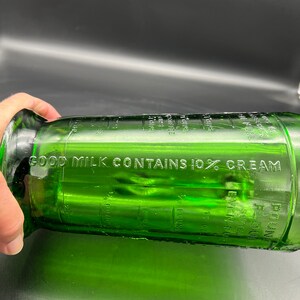 May include: A green glass bottle with the text "GOOD MILK CONTAINS 10% CREAM" etched on the side. The bottle has measurement markings in ounces and pounds. The bottle is cylindrical with a flared base and a narrow neck.