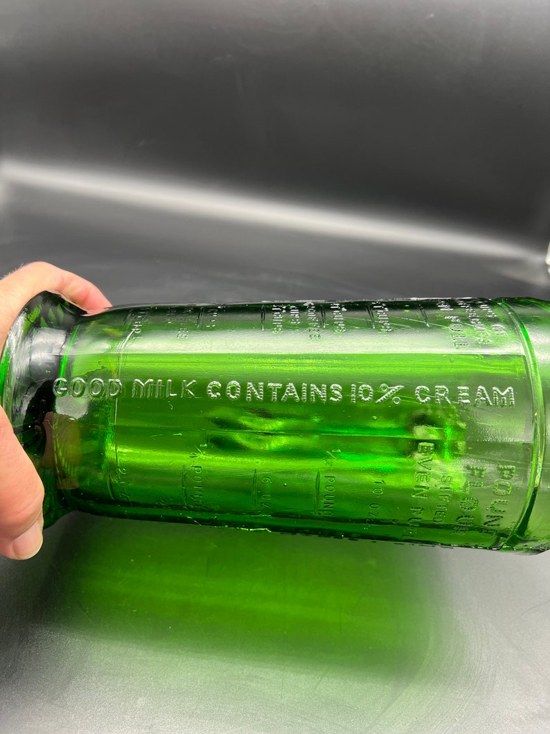 May include: A green glass bottle with the text "GOOD MILK CONTAINS 10% CREAM" etched on the side. The bottle has measurement markings in ounces and pounds. The bottle is cylindrical with a flared base and a narrow neck.