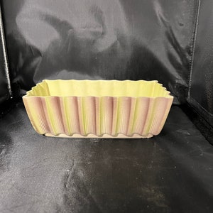 May include: A rectangular ceramic planter with a scalloped edge. The planter has a light yellow interior and a two-tone exterior with vertical stripes of light green and mauve. The planter is designed for indoor or outdoor use.