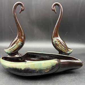 May include: A set of three ceramic swan figurines. Two swans stand upright, featuring a dark brown glaze with green and gold accents. A long, boat-shaped planter with similar colors sits in front.