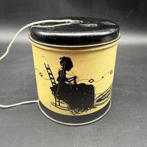 May include: A vintage metal tin with a black lid and a yellow body. The tin features a black silhouette of a person knitting, with a ladder and other decorative elements. A white string is attached to the top.