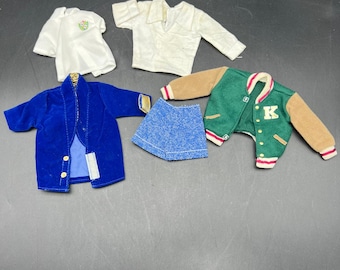 May include: A collection of doll clothes, including a white t-shirt with a green design, a white collared shirt, a blue velvet jacket, denim shorts, and a green and tan letterman jacket with a "K" on it.
