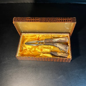 May include: A vintage nutcracker set in a decorative box. The set includes a nutcracker, a pick, and a knife, all with antler handles and silver-toned metal accents. The box is lined with gold fabric and has a brown and orange patterned exterior.