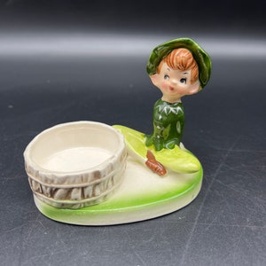 May include: A vintage ceramic planter featuring a seated elf figure with a green hat and outfit. The elf is winking and has red hair. The planter includes a small barrel-shaped container and a light green base with a white interior.