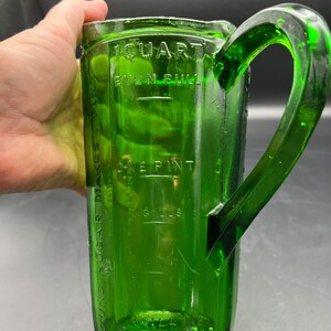 May include: A vintage green glass measuring pitcher with a large handle. The pitcher has measurement markings for quarts, pints, and gills. The pitcher is made of thick, durable glass and has a classic design. The pitcher is a kitchenware item.